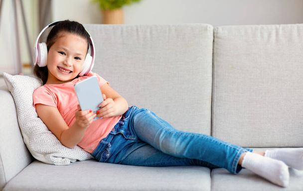 Asian Little Girl In Headphones Using Mobile Phone At Home - Foto, Imagem