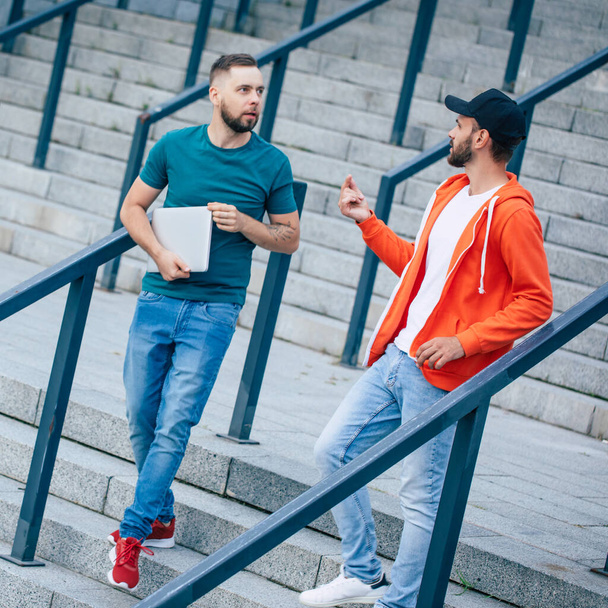Two young friends in casual clothes with smartphones and laptop on the stairs outdoors - Fotoğraf, Görsel
