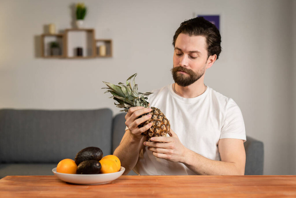 Un jeune homme s'assied à une table et parle de l'utilité des fruits, il les ramasse et montre de l'ananas. Aliments sains - Photo, image