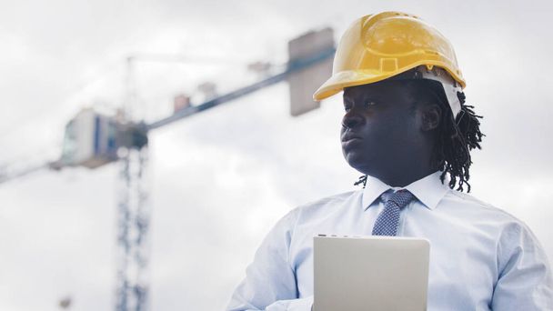 Portrét afrického muže s pevnou čepicí, který používá tablet na staveništi a sleduje práci - Fotografie, Obrázek
