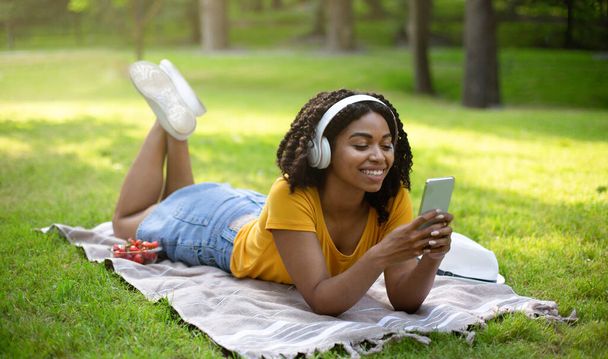 Gelukkige Afro-Amerikaanse vrouw met mobiele telefoon luisteren naar muziek of audio boek in hoofdtelefoon in park - Foto, afbeelding
