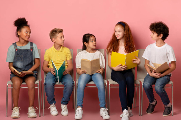 Classmates of different races with school supplies looking at each other on chairs over pink background - Photo, Image