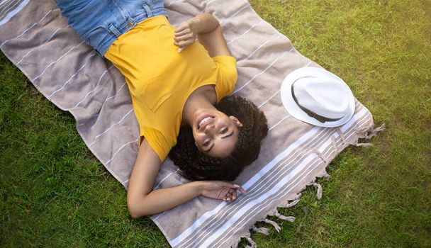 Κάτοψη του ευτυχισμένου μαύρου κοριτσιού χαλαρώνοντας στην κουβέρτα πικνίκ στο πάρκο - Φωτογραφία, εικόνα