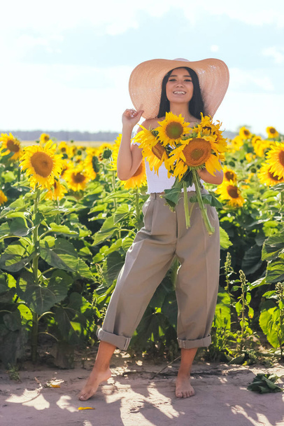 A beautiful brunette woman in a white top on a field with sunflowers holds flowers of sunflowers in her hands. Yellow sunflowers. - Zdjęcie, obraz
