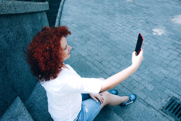 Een meisje met krullend rood haar op de trap, neemt een selfie op een mobiele telefoon. Heldere vrouw buiten het huis. Het gewone leven van een jongedame. Stad levensstijl - Foto, afbeelding