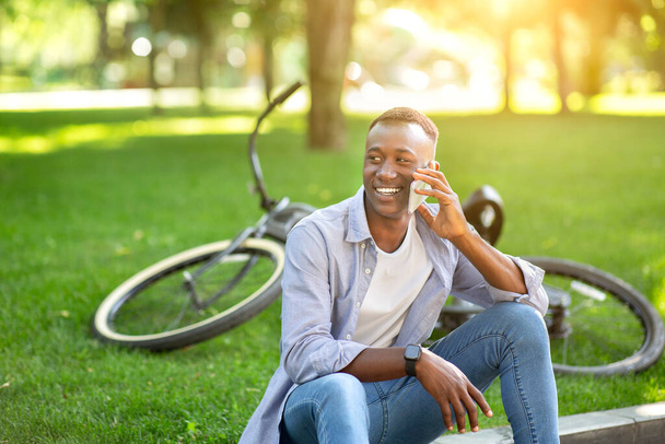 Atrakcyjny czarny mężczyzna siedzi na ziemi koło roweru i rozmawia przez komórkę w parku - Zdjęcie, obraz