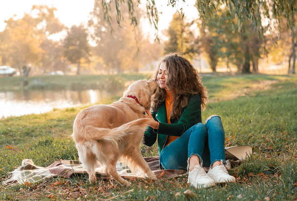 Улыбающаяся женщина обнимает свою собаку-золотистого ретривера у лица. Золотистая собака-ретривер играет с кудрявой женщиной, гуляющей на улице в солнечный день. любовь и забота о питомце. - Фото, изображение