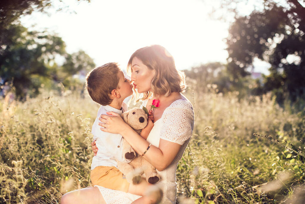 Beautiful, young mom kissing her son on the lips. Boy holding a rose for his mom. Outdoor, nature photo. Mothers day - Foto, Bild