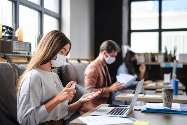 Portrait of young businesspeople with face masks working indoors in office, disinfecting hands. - Foto, Bild