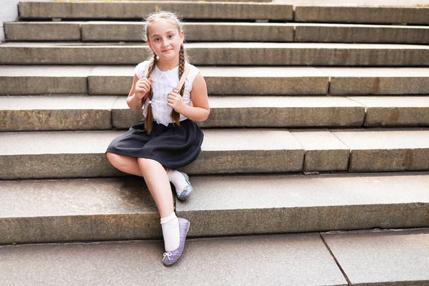 милая маленькая школьница с рюкзаком сидит на большой каменной лестнице в теплый день - Фото, изображение