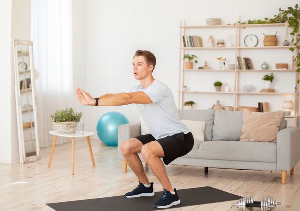 Keeps fit at home. Young man in sportswear and with fitness tracker squats in interior - Foto, immagini