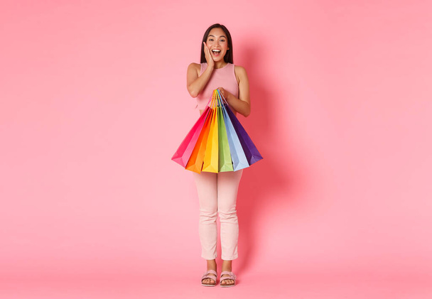 Ganztägiges Porträt eines amüsierten und glücklichen, fröhlichen asiatischen Mädchens, das die Wange berührt und lächelt, während es Einkaufstüten hält, viele Dinge mit Rabatten, Verkäufen, stehendem rosa Hintergrund kauft - Foto, Bild