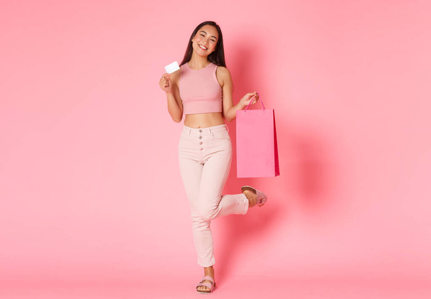 Повна тривалість безтурботних і мрійливих, мила азіатська дівчина позує з кредитною карткою і сумками на рожевому тлі, насолоджуючись вихідними в торгових центрах, оточених спеціальними пропозиціями і знижками
 - Фото, зображення