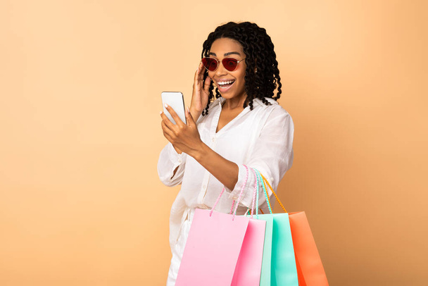 Dame noire excitée utilisant des sacs à dos de téléphone Holding Shopper, fond beige - Photo, image