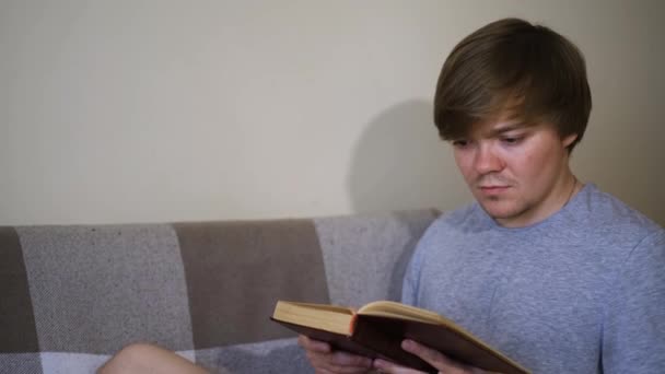 若い男はソファーに座って本を熱心に読んでいる。コンセプト。学生は緊張した表情で古典を読む。男は古い本を読む。旧古典文学 - 映像、動画
