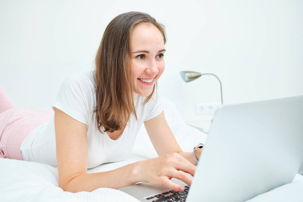 Egy fehér nő fekszik az ágyon, laptopot használ, hogy megnézze.. - Fotó, kép