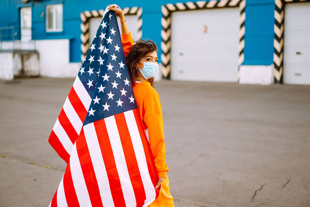 African American woman with medical face mask posing with american flag. Curly woman during quarantine. The concept of preventing the spread of the epidemic. Covid-2019. - Foto, Imagem