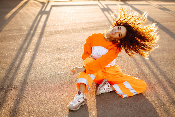 Portrait of stylish woman with curly hair in an orange suit. Young African American woman hip-hop dancer at sunset. Sport, dancing and urban culture concept.  - Фото, изображение