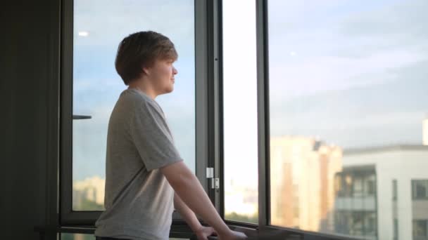 A fiatalember élvezi a kilátást az erkélyen álló ablakból. Fogantatás. A diák boldogan nézi az életet az ablakon túl a háttér égen. Diák áll az erkélyen, és néz ki az ablakon - Felvétel, videó