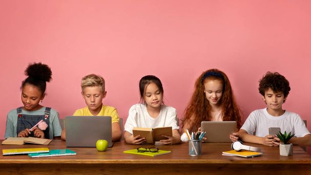 Enfants multiethniques avec appareils électroniques et livres étudiant à table sur fond rose, espace libre - Photo, image