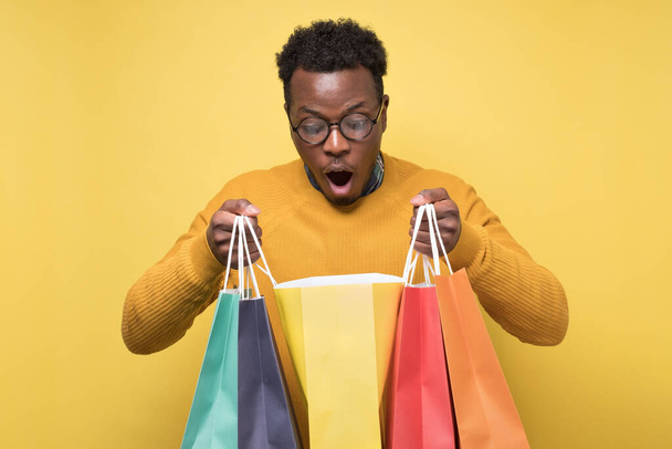 Shocked african young man holding shopping bags being surprises. Wow, what I have bought. Studio shot on yellow wall. - Foto, imagen