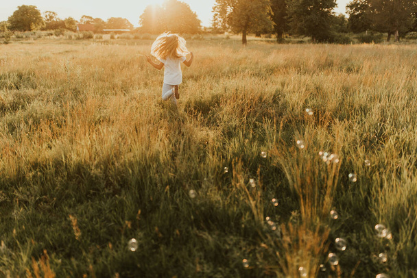 Ένα μικρό ξανθό κορίτσι ντυμένο με ανοιχτόχρωμα ρούχα και ένα λευκό μπλουζάκι τρέχει μακριά από σαπουνόφουσκα. Κατά μήκος του ψηλού πράσινου χόρτου προς τον ήλιο που δύει. Ζεστό καλοκαίρι ηλιόλουστο καιρό στο ηλιοβασίλεμα. - Φωτογραφία, εικόνα