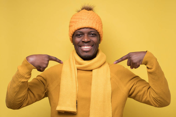 Sonriente, joven africano con suéter cálido, bufanda y sombrero señalando con los dedos en el pecho. Seleccioname por favor. Emoción humana positiva - Foto, Imagen