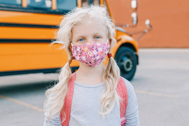 Een blanke studente met gezichtsmasker in de buurt van een gele bus. Kind met persoonlijke beschermingsmiddelen op het gezicht. Onderwijs en terug naar school in september. Nieuw normaal tijdens het coronavirus.  - Foto, afbeelding