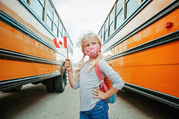 カナダの国旗を掲揚したフェイスマスクをした白人女子学生。カナダの黄色のスクールバスの近くの学生の子供。教育と9月に学校に戻る。コロナウイルス中の新しい正常.  - 写真・画像