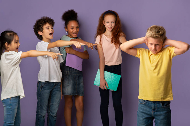 Prevention of bullying. Depressed boy closing ears while his classmates laughing at him over lilac background - Foto, afbeelding