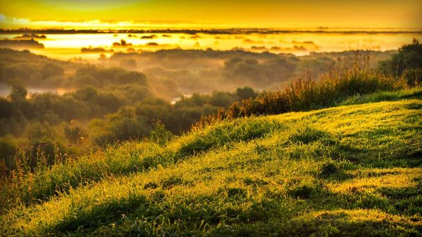 Piękny widok ze szczytu wzgórza z zieloną trawą pokrytą kroplami rosy do mglistej doliny w świetle wschodzącego słońca - Zdjęcie, obraz