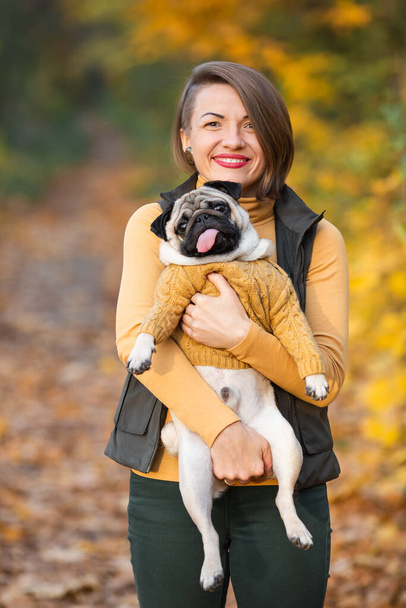 Dziewczyna trzyma swojego psa mopsa jesienią w parku. - Zdjęcie, obraz