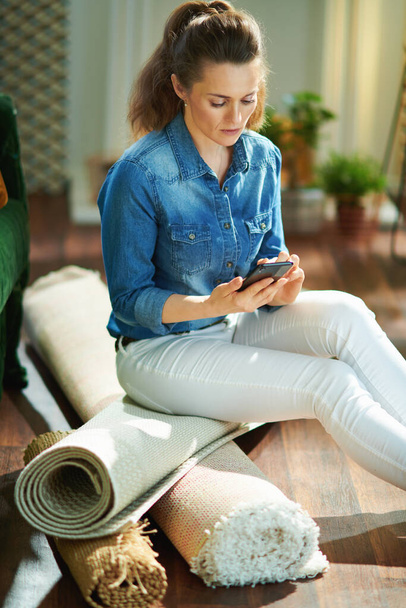pensive young woman in jeans shirt and white pants with pile of carpets searching for carpet cleaning service on a smartphone in the modern living room in sunny day. - Photo, Image