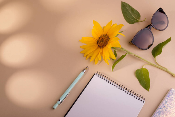 Fashion flat lag met notebook, zonnebril, zonnebloem, en pen tegen beige achtergrond. Goed voor reclame in de zomer. - Foto, afbeelding