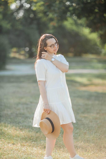 Νεαρή γυναίκα πορτρέτο σε ηλιαχτίδες. Μια όμορφη νεαρή γυναίκα σε ένα λευκό φόρεμα με ψάθινο ψάθινο καπέλο περπατά στο πάρκο. - Φωτογραφία, εικόνα