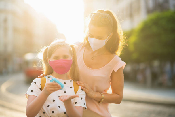 Life during coronavirus pandemic. young mother and school child with masks and yellow backpack disinfecting hands with sanitiser and getting ready for school outdoors. - Photo, Image
