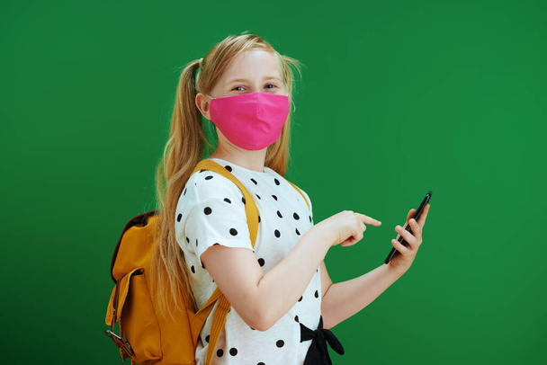 Life during coronavirus pandemic. modern pupil in white polka dot blouse with yellow backpack and pink mask using smartphone applications against chalkboard green background. - Fotoğraf, Görsel