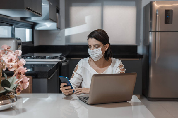 Mladá žena pracující ve své domácí kanceláři, se svým notebookem, a nosí ochrannou masku proti viru koróny, covid 19 - Fotografie, Obrázek