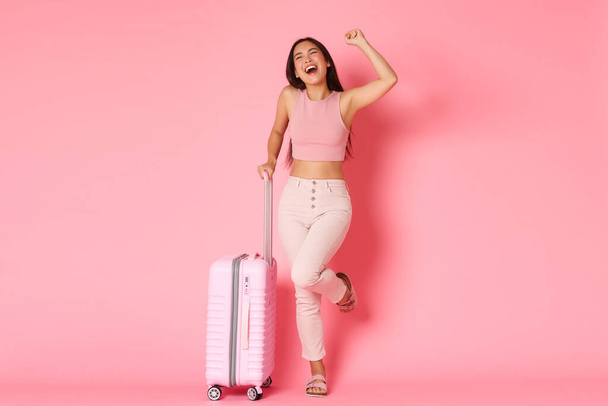 Travelling, holidays and vacation concept. Excited and happy smiling asian girl, screaming yes and fist pump in air as celebrating start of adventures to paradise summer resort, hold suitcase - Foto, Imagen