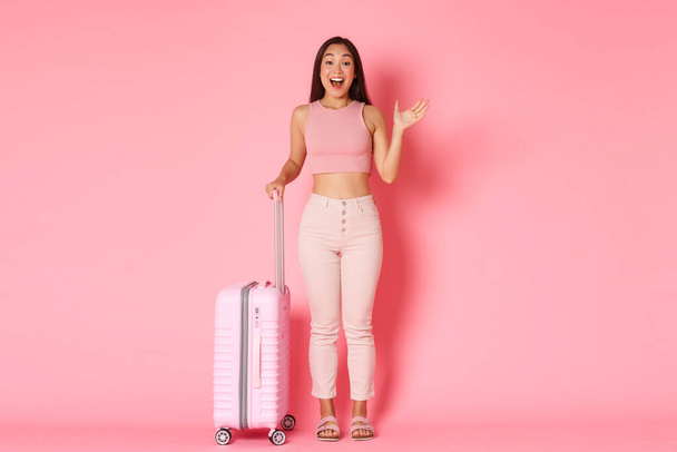Reisen, Urlaub und Urlaubskonzept. Fröhliches Glamour-asiatisches Mädchen in Sommerkleidung, Touristin winkt in Hallo-Geste, als jemand ihren Flughafen trifft, Koffer über rosa Hintergrund haltend - Foto, Bild