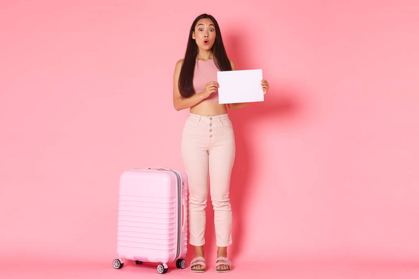 Путешествие, отпуск и концепция отпуска. Полная длина забавной и очаровательной азиатской красивой девушки турист стоит с листом бумаги, подписать над розовым фоном с чемоданом - Фото, изображение