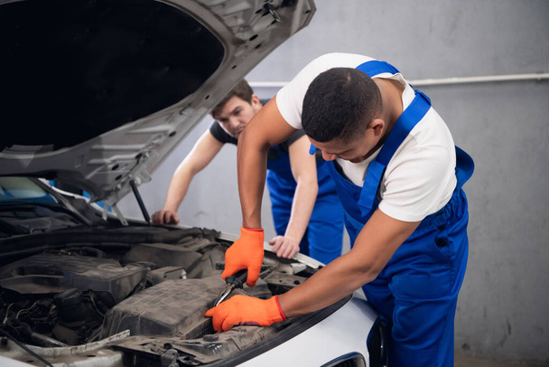Dos mecánicos masculinos en uniforme inspeccionan un motor de coche - Foto, Imagen