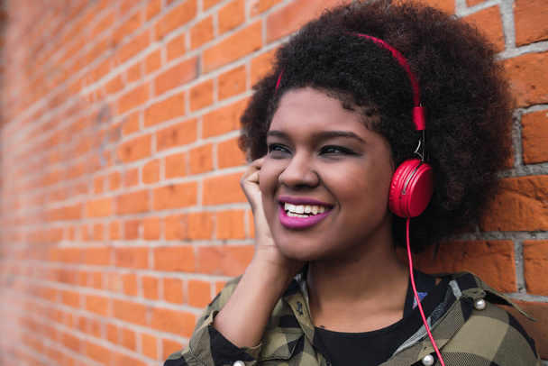 Portrait of Afro american latin woman smiling and listening to music with headphones against brick wall. Outdoors. - Photo, Image
