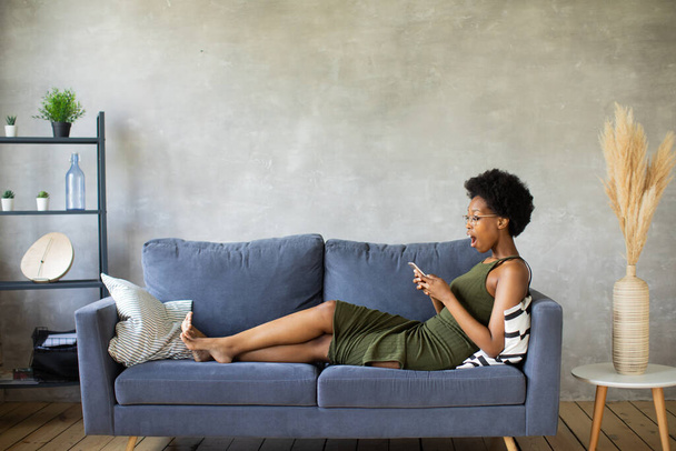 Happy african American young woman sit relax on cozy couch happy to move to new apartment. Smiling black girl rest on comfortable sofa in living room dreaming - Фото, зображення
