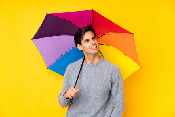 Άντρας κρατώντας μια ομπρέλα πάνω από απομονωμένο κίτρινο φόντο γελώντας και κοιτώντας ψηλά - Φωτογραφία, εικόνα