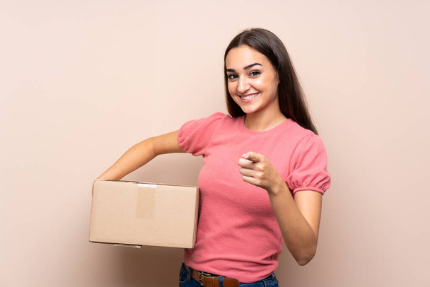Молода жінка над ізольованим тлом тримає коробку, щоб перенести її на інший сайт, вказуючи на передній план
 - Фото, зображення