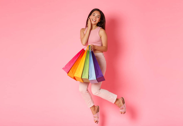 Full length portrait of gorgeous dreamy and carefree asian female tourist enjoying shopping in foreign malls, jumping cheerfully and holding shop bags, posing over pink background - Photo, image