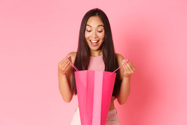 Beauty, Mode und Lifestyle-Konzept. Nahaufnahme eines aufgeregten und erstaunten, überraschten Mädchens, das in einer Einkaufstasche mit fröhlichem, optimistischem Lächeln vor rosa Hintergrund steht - Foto, Bild