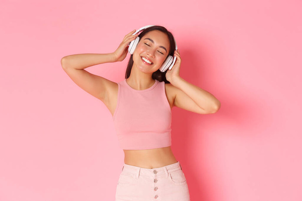 Despreocupado hermosa chica asiática en auriculares, bailando con los ojos cerrados y relajados, sonrisa feliz, disfrutando de escuchar música, escuchar canción favorita, de pie sobre fondo rosa - Foto, imagen