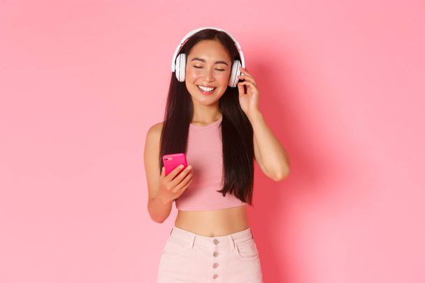 Портрет привлекательной женщины, которая наслаждается любимой песней, закрывает глаза, чтобы расслабиться, слушая музыку в наушниках, держа мобильный телефон и стоя на розовом фоне - Фото, изображение